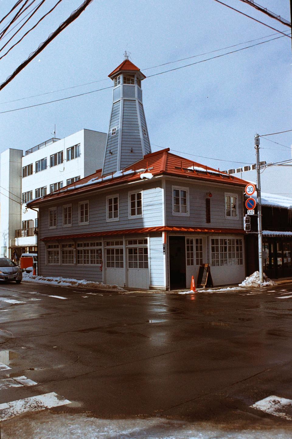 A light blue building with a red roof and a tall, narrow tower stands on a street corner with patches of snow on the ground.