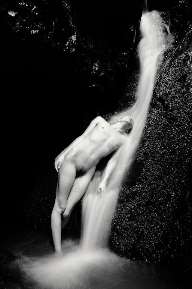 A nude man stands under a waterfall in a dark grotto.