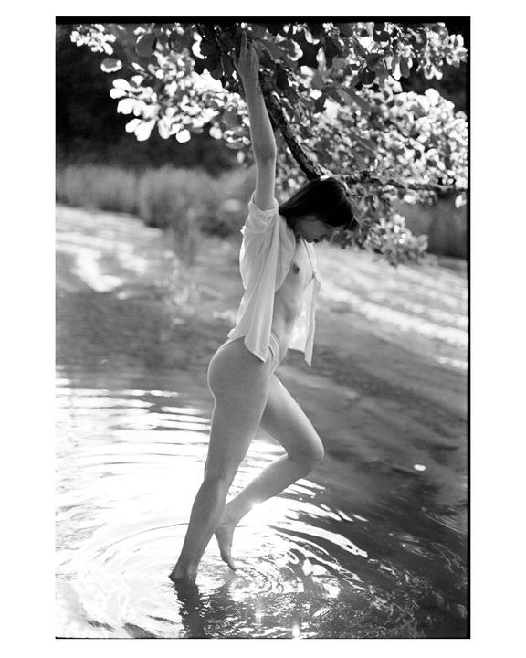 A woman stands in water, holding onto a tree branch, with sunlight reflecting on the water.