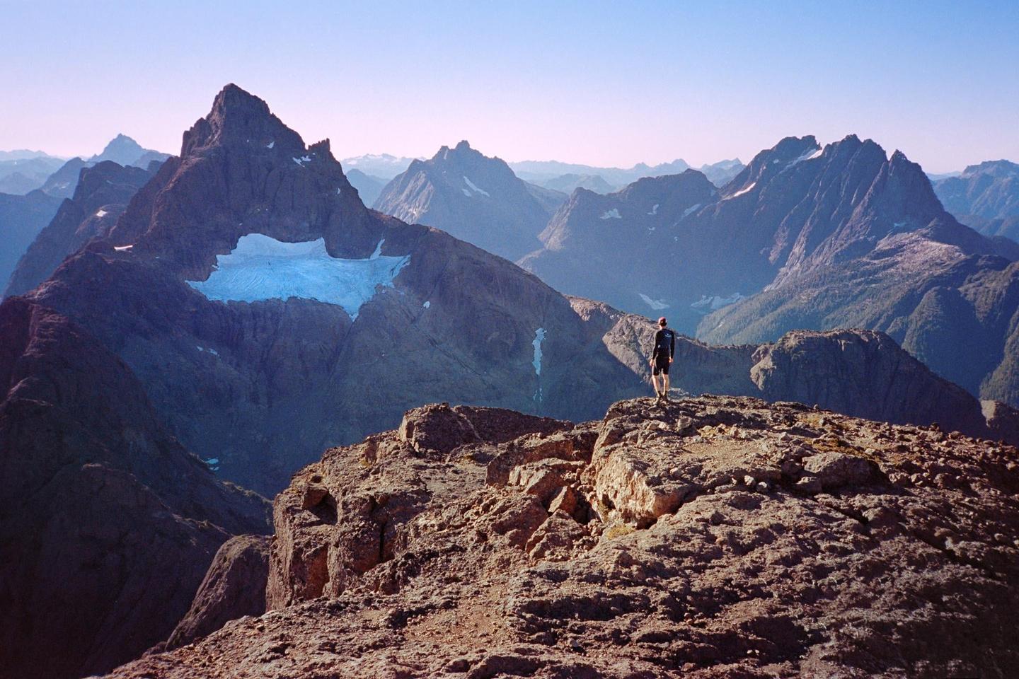 A man stands on a rocky mountain peak overlooking a vast mountain range with a glacier.
