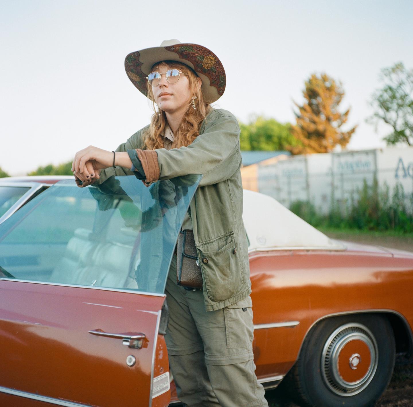 A woman wearing a cowboy hat and sunglasses leans on the open door of a vintage car.