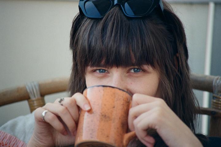 A woman with bangs and sunglasses on her head holds an orange mug to her mouth, looking directly at the viewer.