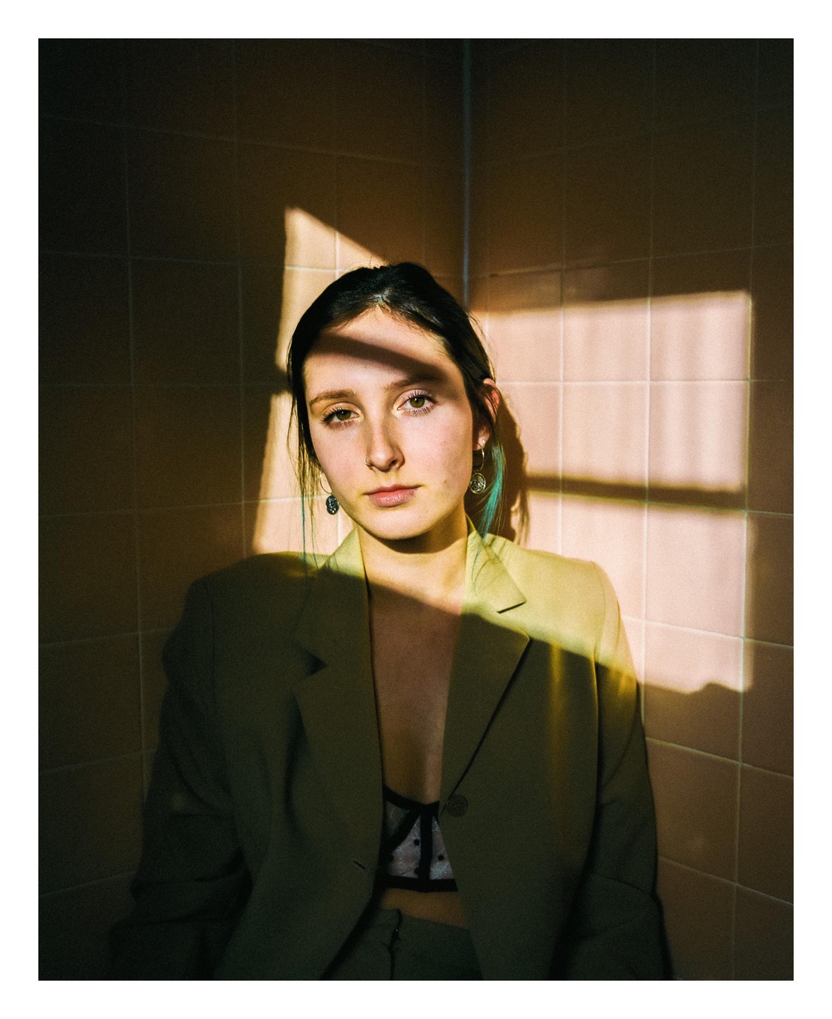 A woman wearing a green suit jacket and a bra sits in a corner with window light and shadow falling on her.