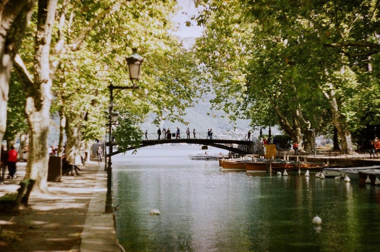 People walk across a bridge over a canal lined with trees and boats.