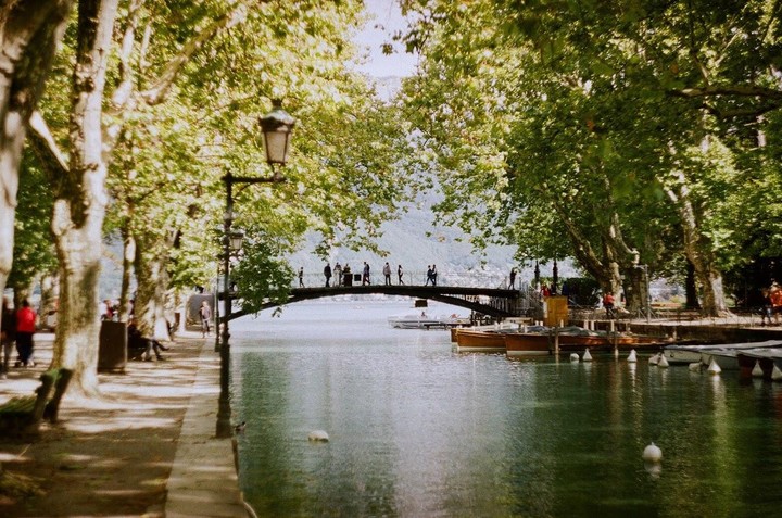 People walk across a bridge over a canal lined with trees and boats.