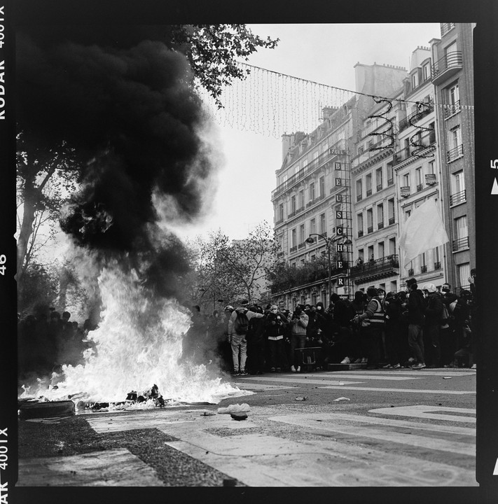 A large fire burns in the street with a crowd of people and buildings in the background.