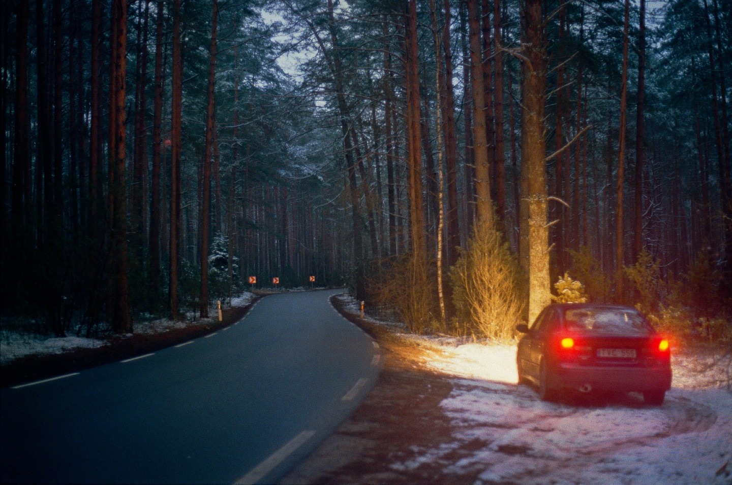 A car with its brake lights on is parked on the snowy shoulder of a winding road through a forest at dusk.