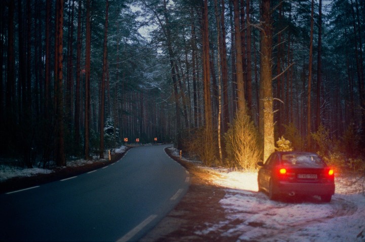 A car with its brake lights on is parked on the snowy shoulder of a winding road through a forest at dusk.