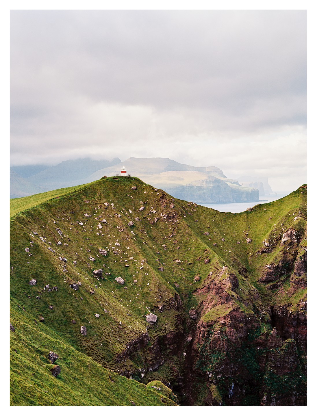 A red and white lighthouse stands on a green, rocky hill overlooking a body of water and distant mountains under a cloudy sky.