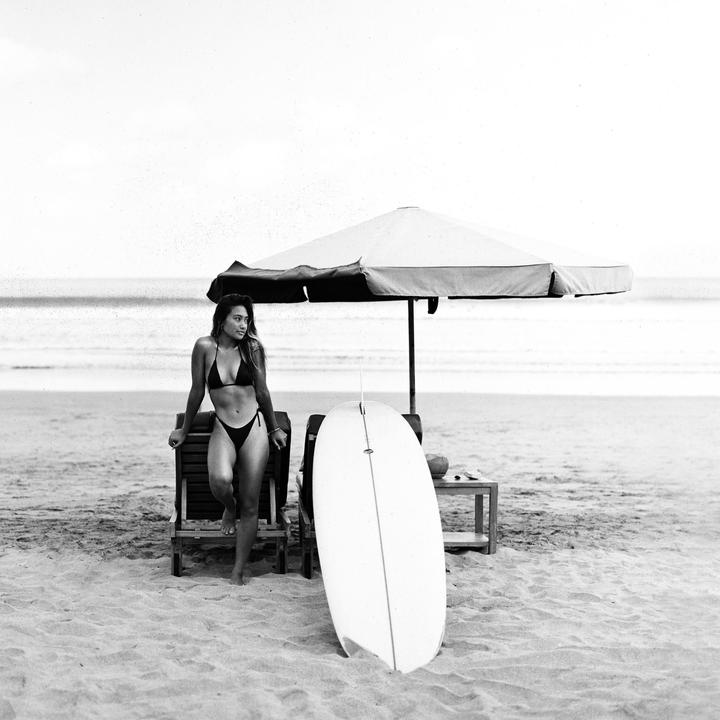 A woman in a bikini stands next to a surfboard and beach chair under an umbrella on a sandy beach.