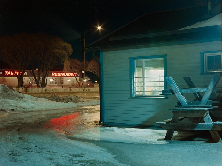 A restaurant with a neon sign is visible across a snowy street from a building with a picnic table turned upside down in front of it.
