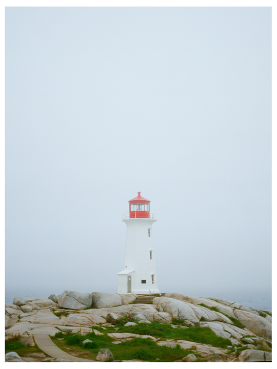 A white lighthouse with a red top stands on a rocky coast under a hazy sky.