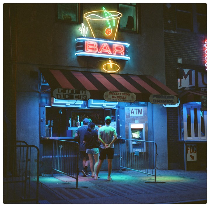 Three people stand in front of a bar with neon signs at night.