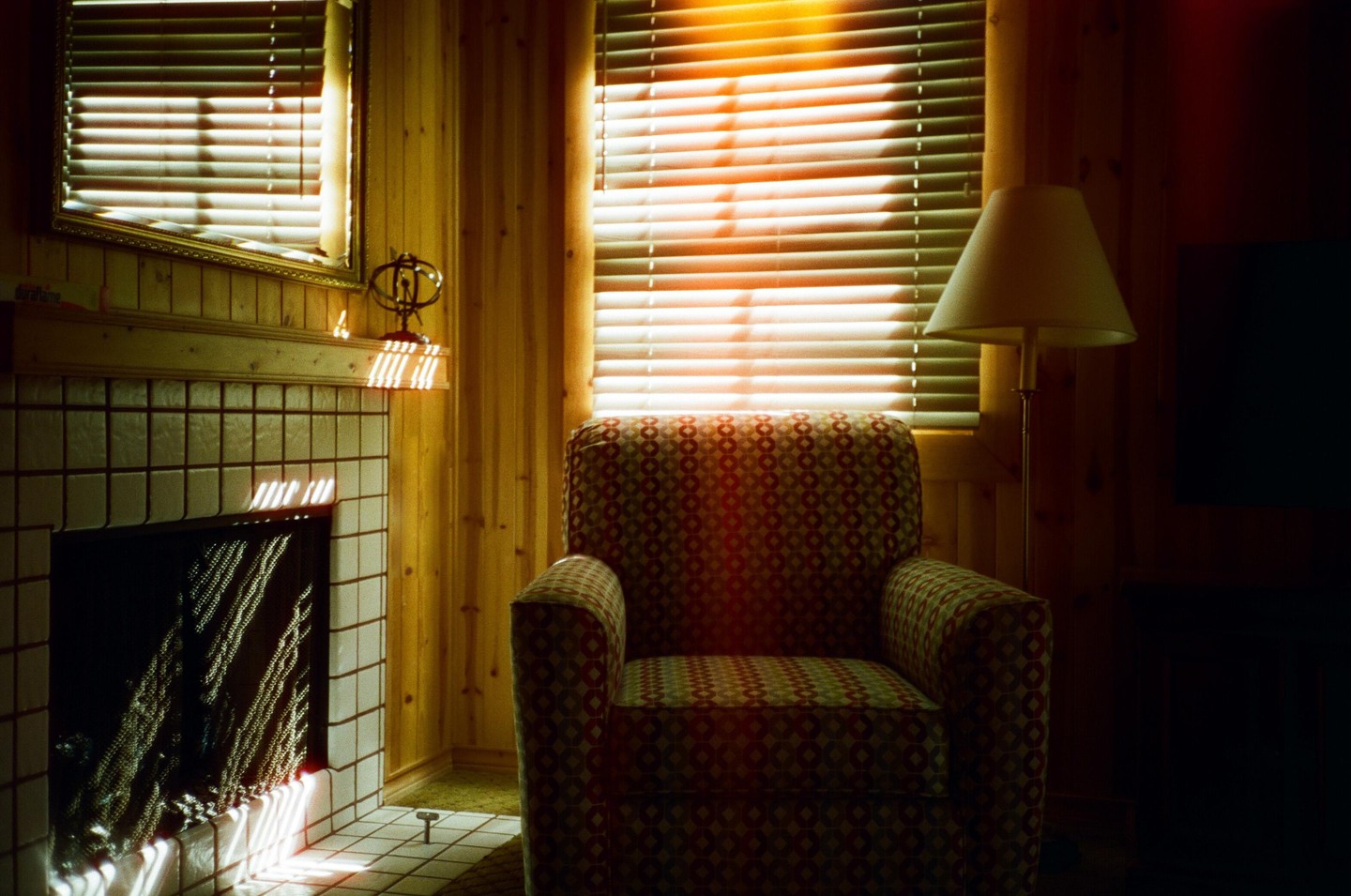 Sunlight streams through blinds onto a patterned armchair and a fireplace in a wood-paneled room.