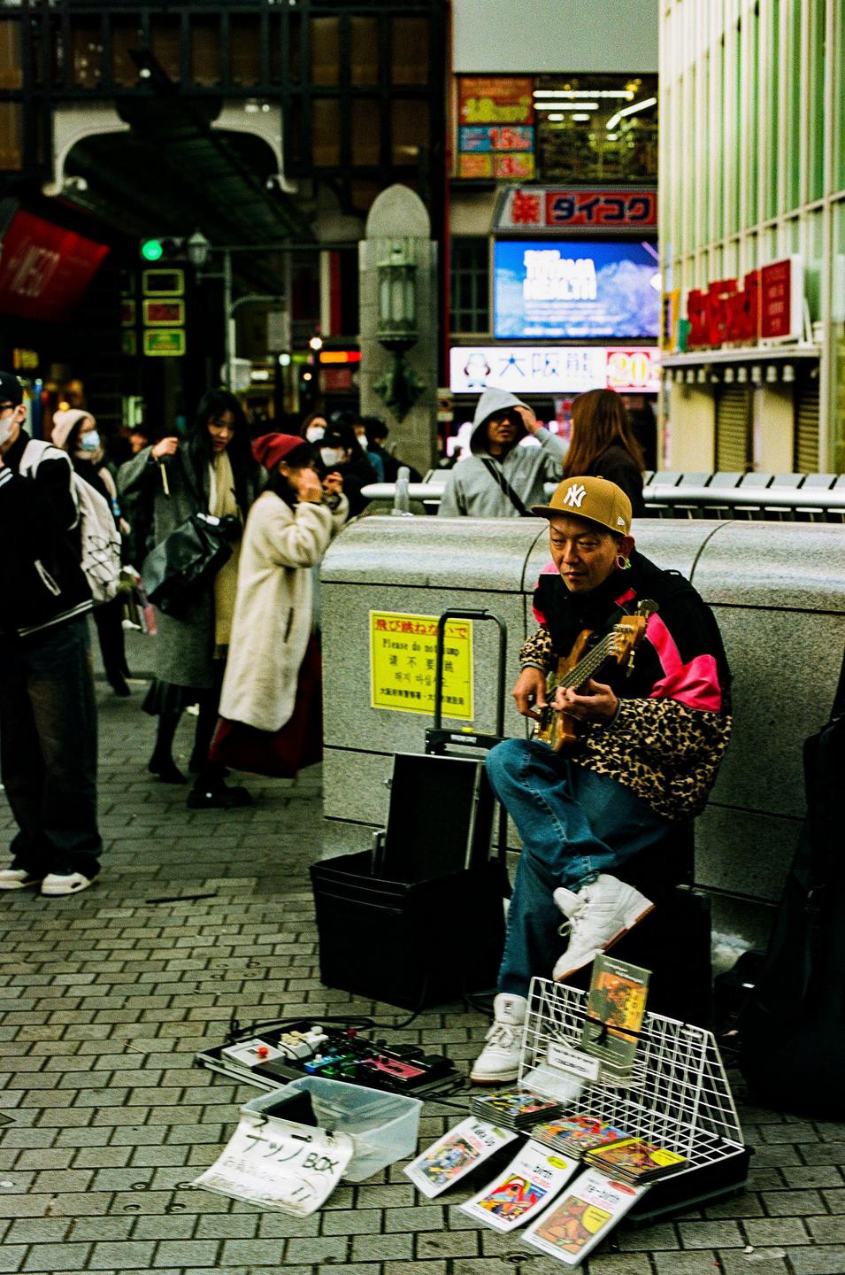 A man wearing a leopard print jacket and a baseball cap plays a ukulele on a city street.