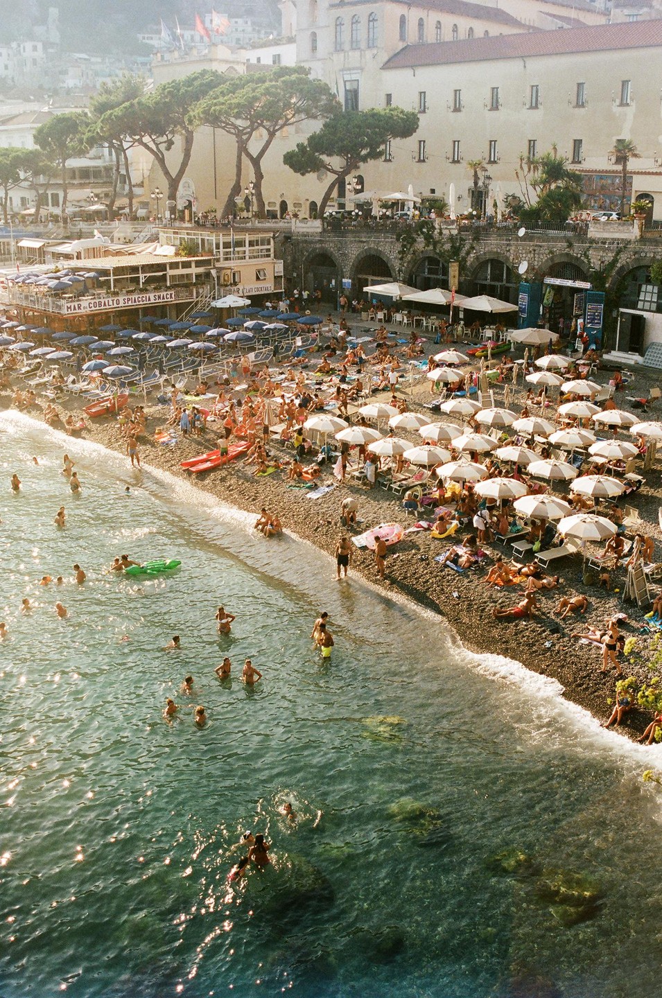A crowded beach with many people swimming in the water and sunbathing under umbrellas on the shore, with buildings and trees in the background.