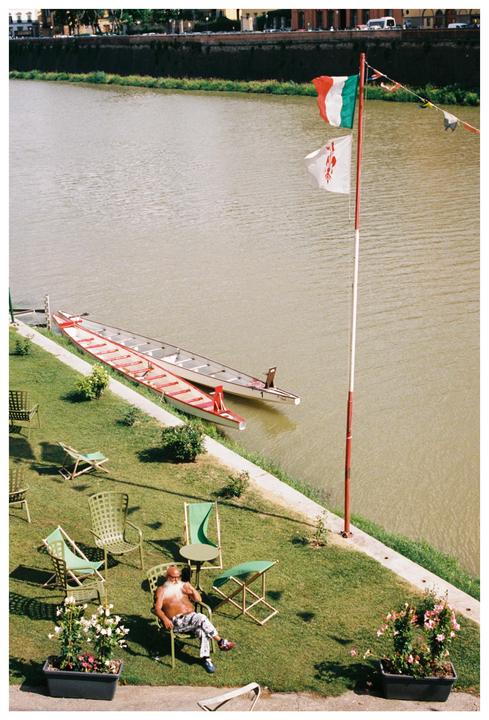 A man with a long white beard sits shirtless in a chair on a grassy bank next to a river with two boats and an Italian flag.
