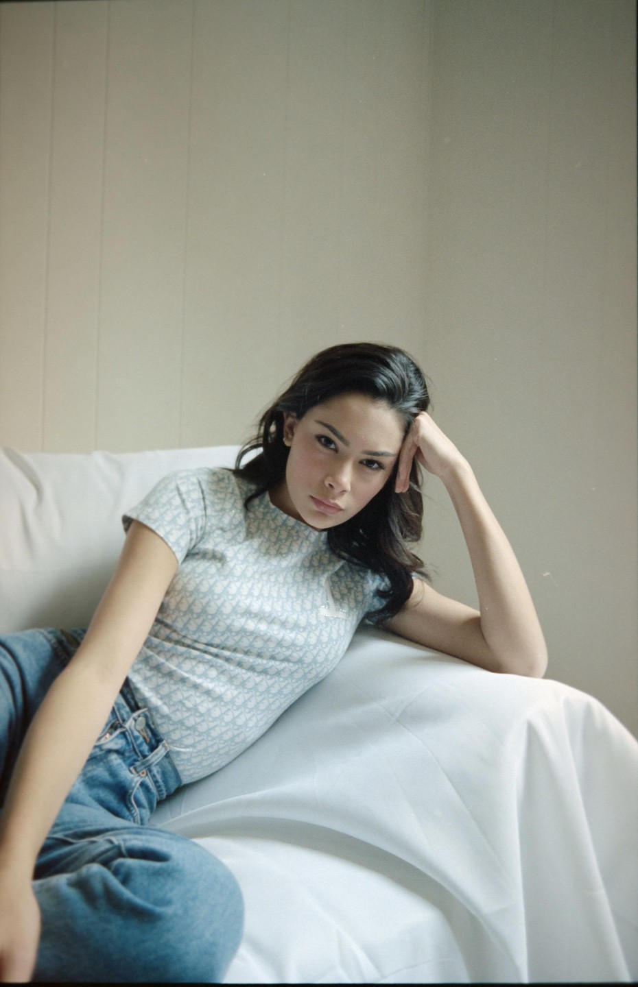 A woman with dark hair reclines on a white sofa, wearing a light blue patterned shirt and jeans, looking directly at the viewer.
