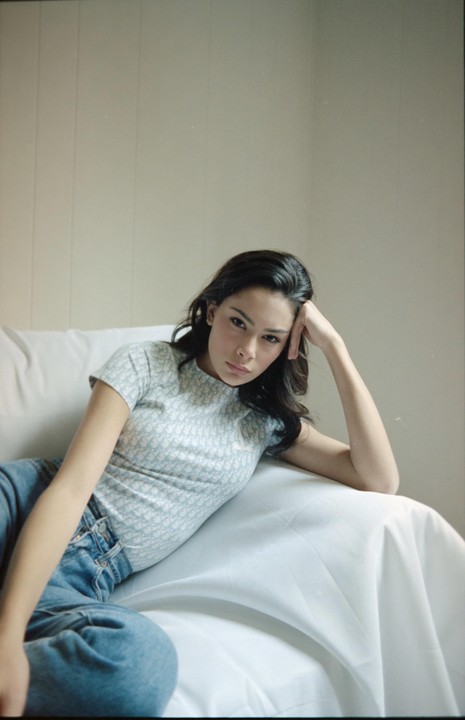 A woman with dark hair reclines on a white sofa, wearing a light blue patterned shirt and jeans, looking directly at the viewer.