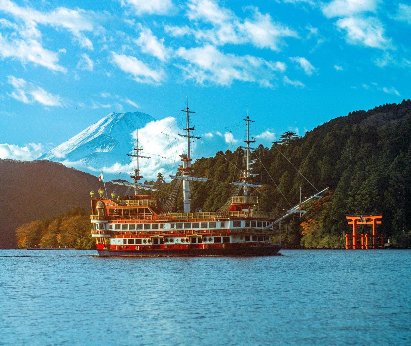 A large boat with multiple masts sails on a lake with a snow-capped mountain and a red torii gate in the background.
