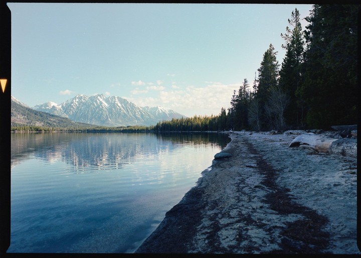 A lake with mountains and a forest in the background is seen from a sandy shore.