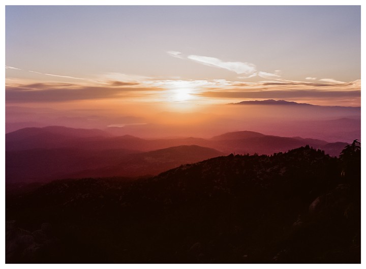 The sun sets over a mountain range with a hazy sky.