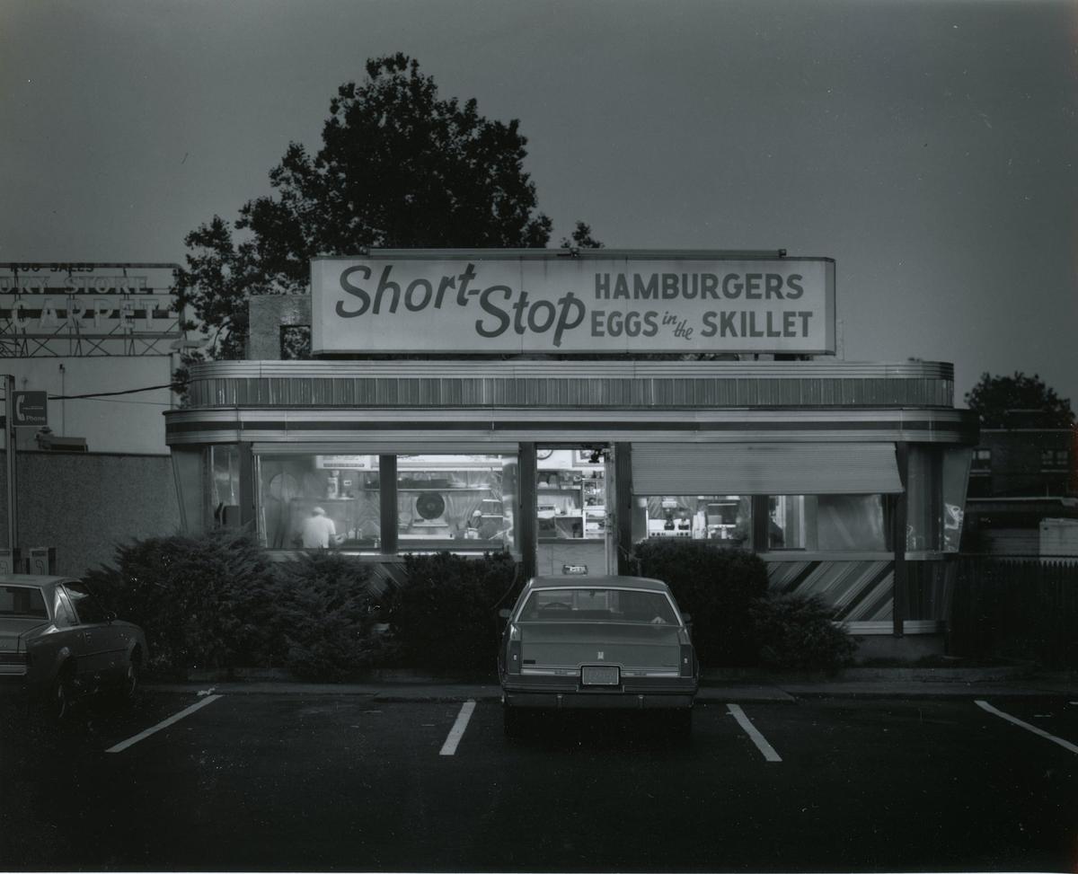 A diner with a brightly lit interior and a sign that reads "Short Stop Hamburgers Eggs in the Skillet" is seen from a parking lot at night.