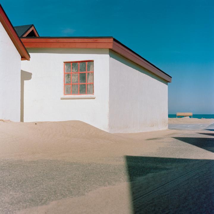 A white building with a red-framed window is partially covered by a sand dune next to a body of water.
