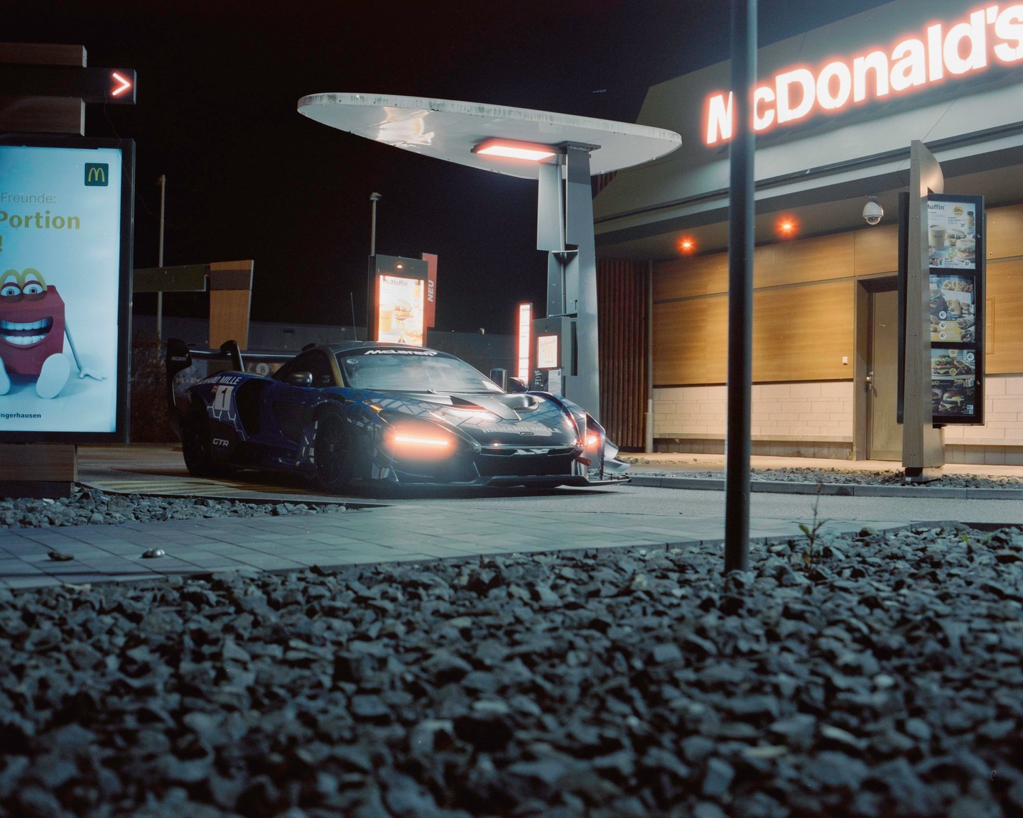 A blue race car with its headlights on is parked at a McDonald's drive-thru at night.