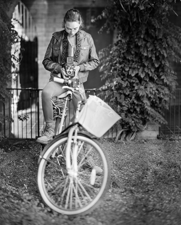 A woman sits on a bicycle and holds a twin lens reflex camera.