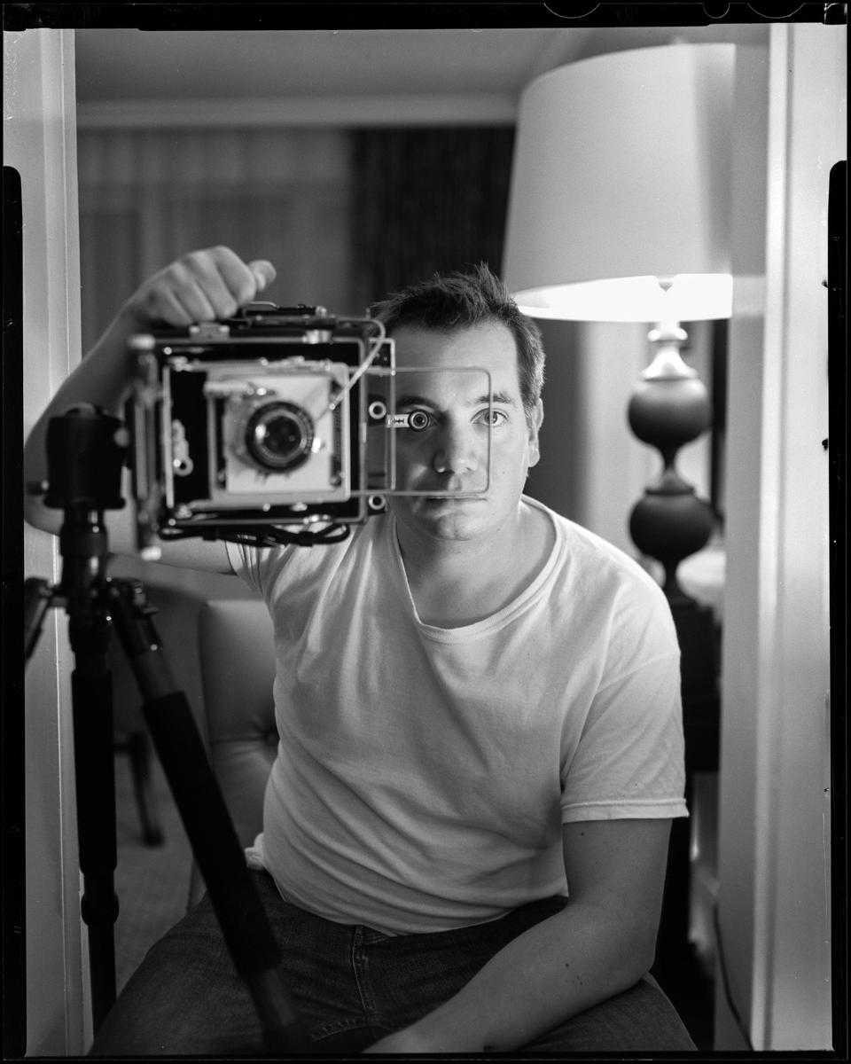 A man holds a large format camera in front of his face, taking a self-portrait in a mirror.