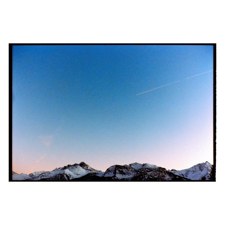 A jet contrail crosses a clear blue sky above a range of snow-capped mountains.