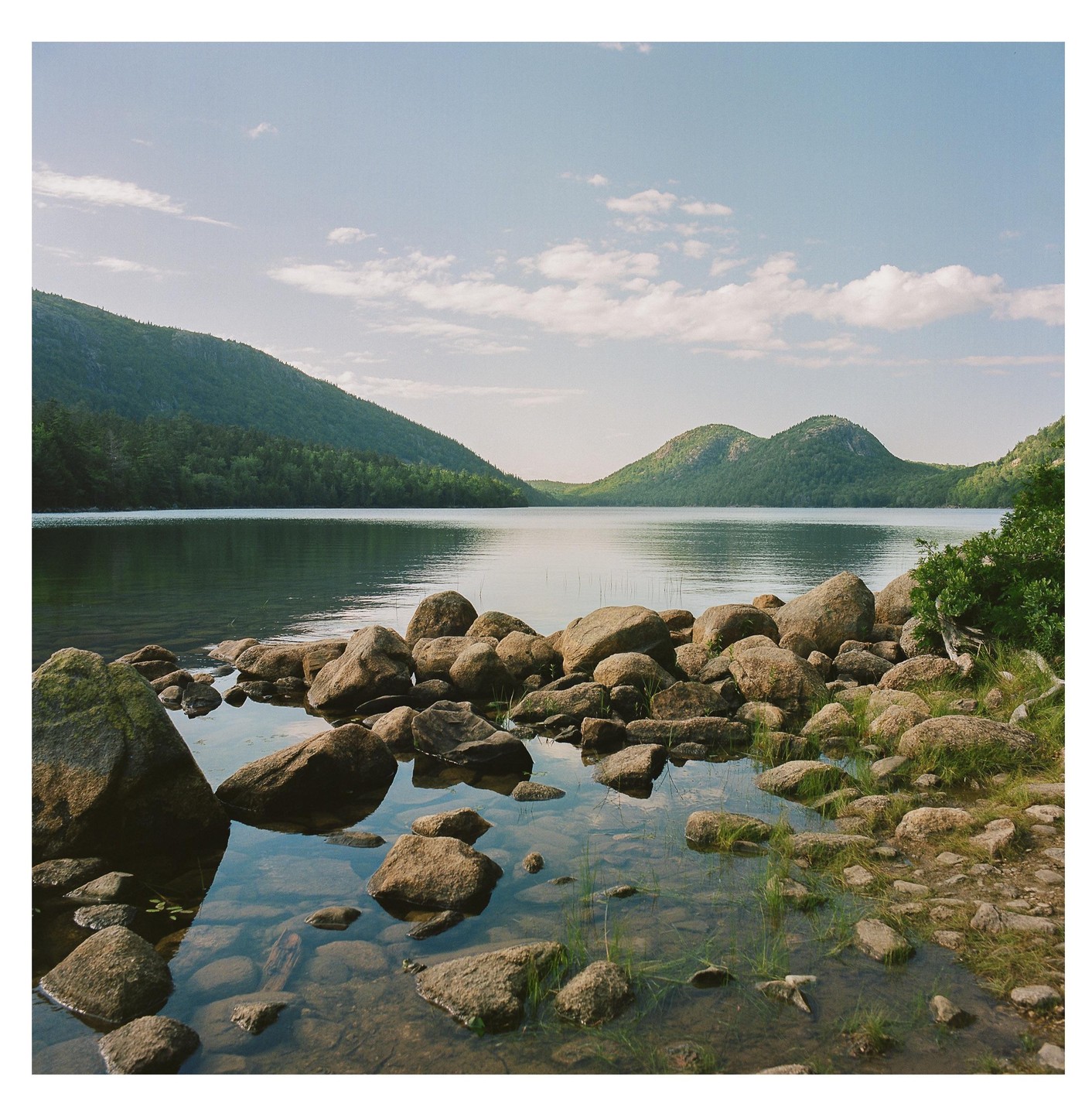 A rocky shore leads into a calm lake surrounded by tree-covered mountains under a partly cloudy sky.