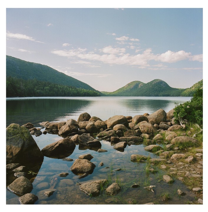 A rocky shore leads into a calm lake surrounded by tree-covered mountains under a partly cloudy sky.