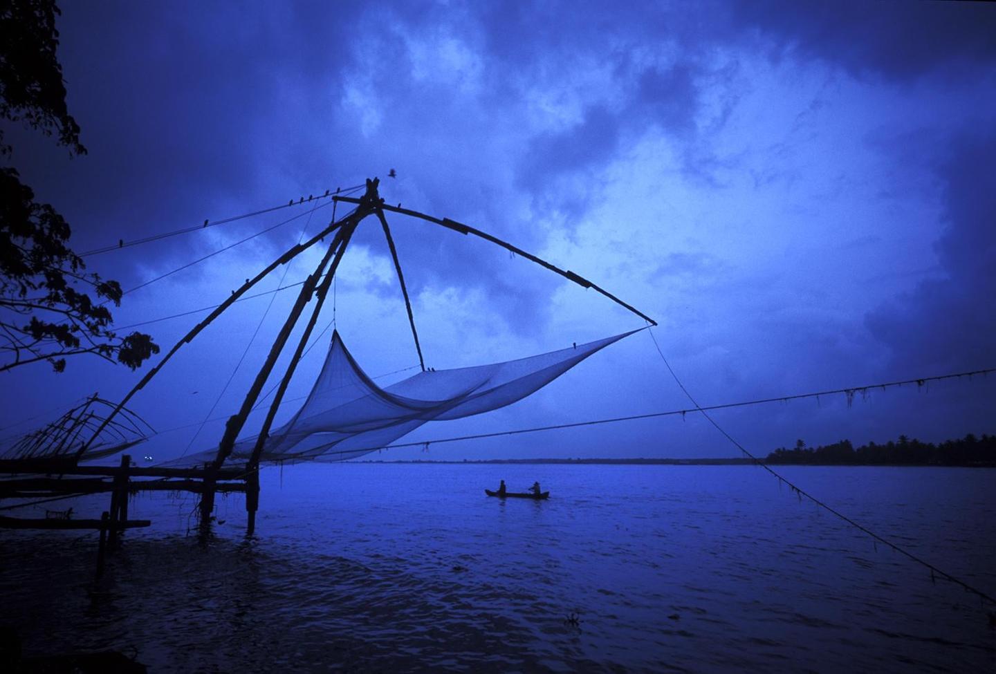A large fishing net structure extends over a body of water with a small boat and two people in the distance under a cloudy blue sky.