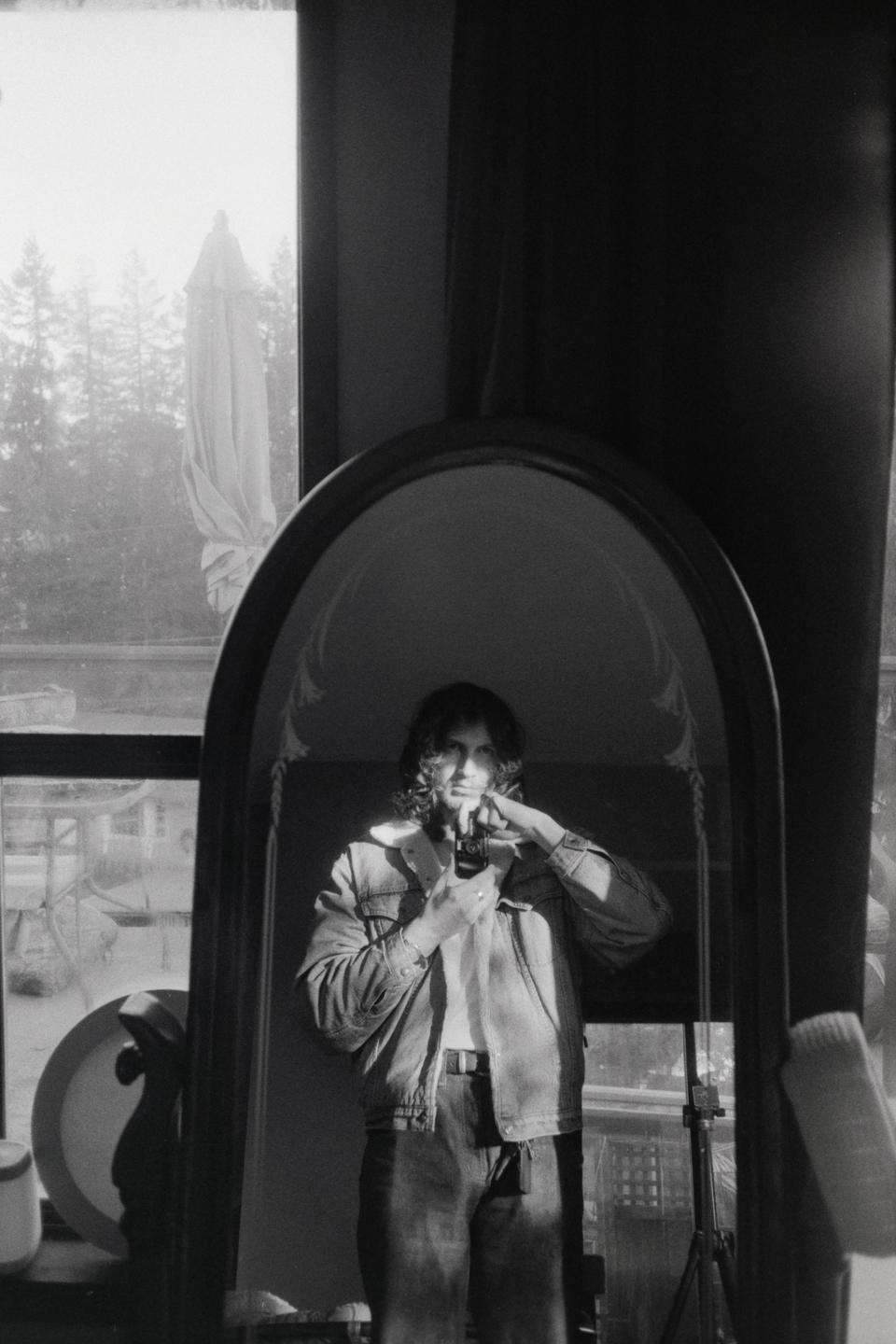 A man with long hair wearing a denim jacket takes a self-portrait in a mirror.