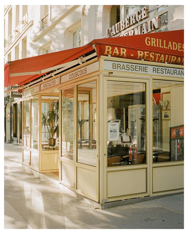 A brasserie with a red awning and glass enclosure stands on a sunlit street.