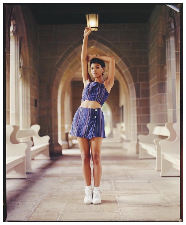 A woman stands with her arms raised in a stone hallway with arched ceilings and benches.