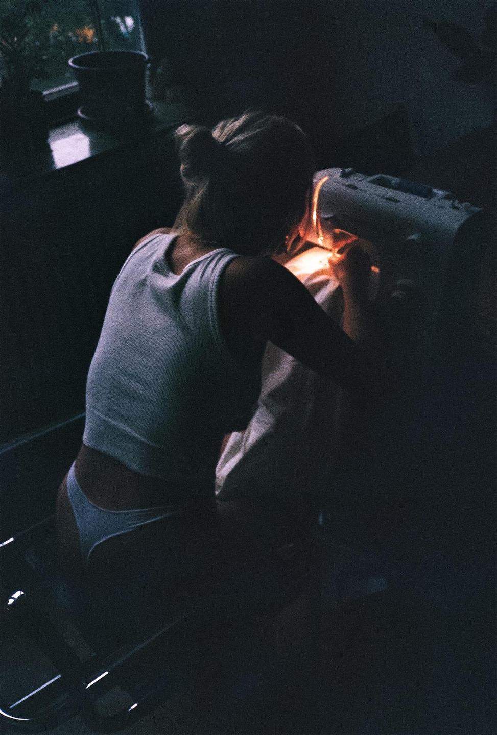 A woman in a tank top and underwear is sewing in a dark room, illuminated by the light of the sewing machine.