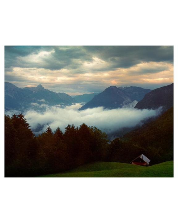 A small house sits on a green hillside overlooking a valley filled with fog and surrounded by mountains under a cloudy sky.