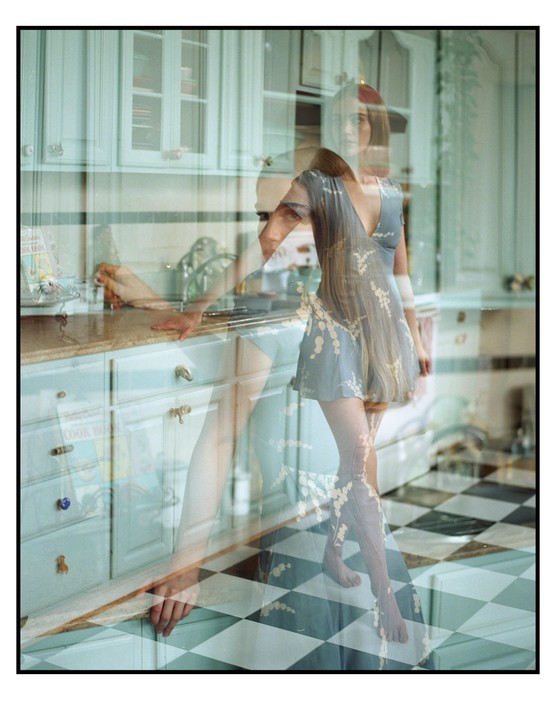 A woman stands in a kitchen with light blue cabinets and a checkered floor, with a double exposure showing her in two positions.
