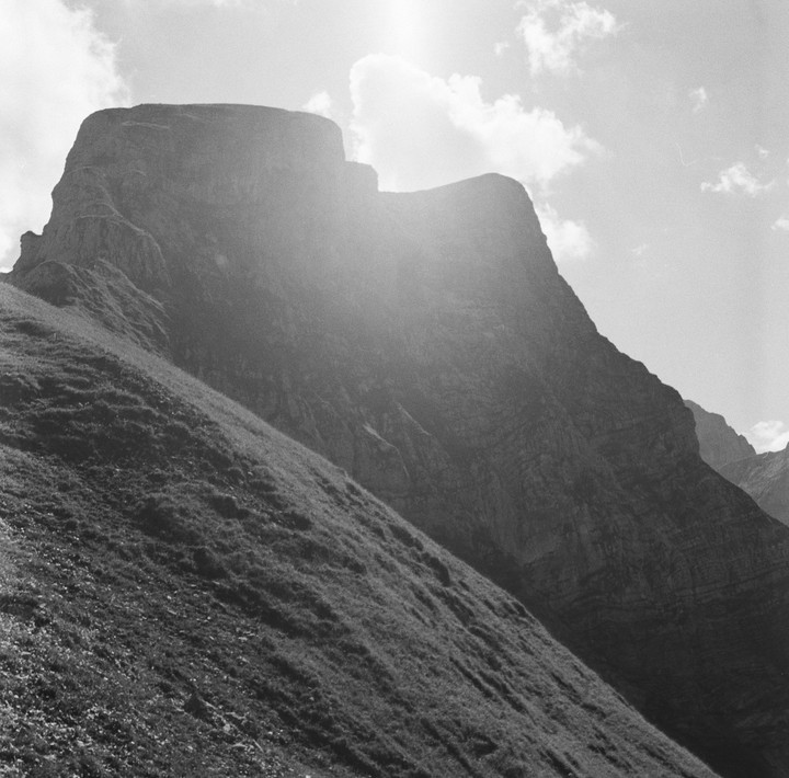A mountain with a grassy slope in the foreground is backlit by the sun.