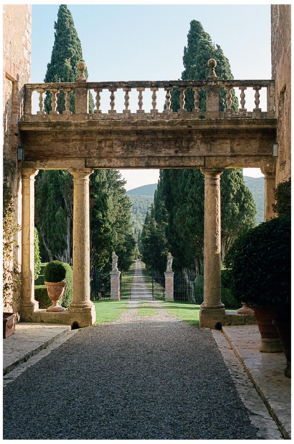 A long gravel driveway leads through a gate and a row of tall trees towards a distant mountain, framed by a stone archway and balcony.