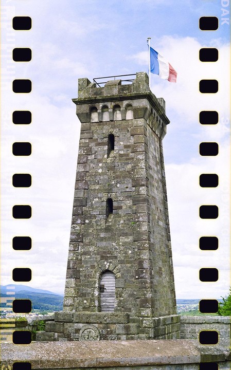A stone tower with a French flag flying from its top stands against a cloudy sky, with a landscape visible in the distance.