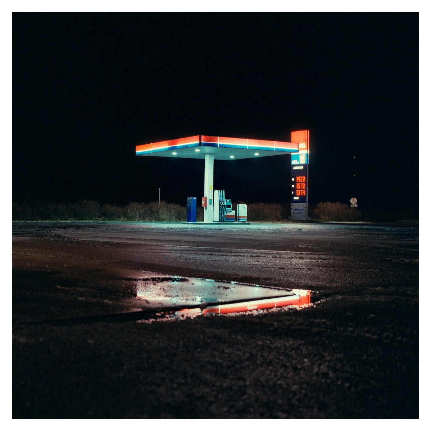 A gas station with a red and blue canopy is illuminated at night, with a puddle reflecting the lights in the foreground.