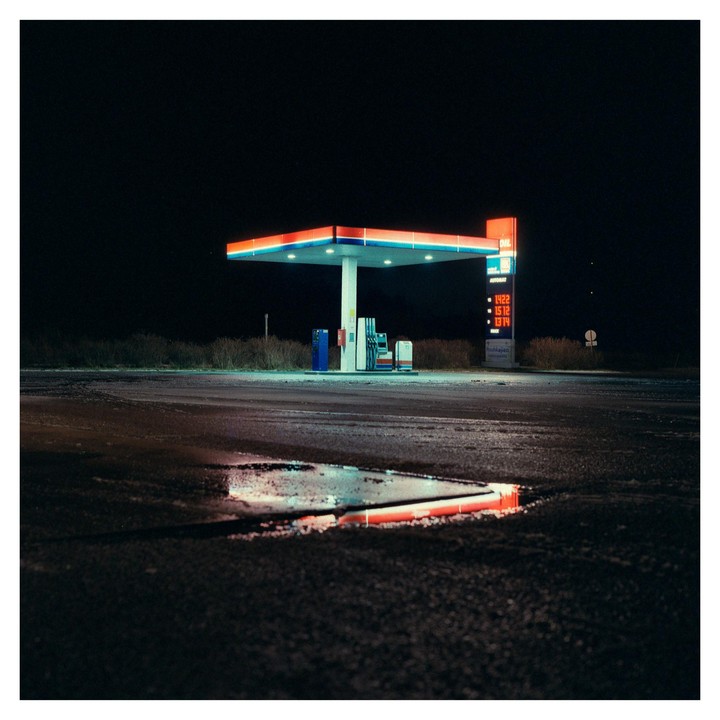A gas station with a red and blue canopy is illuminated at night, with a puddle reflecting the lights in the foreground.