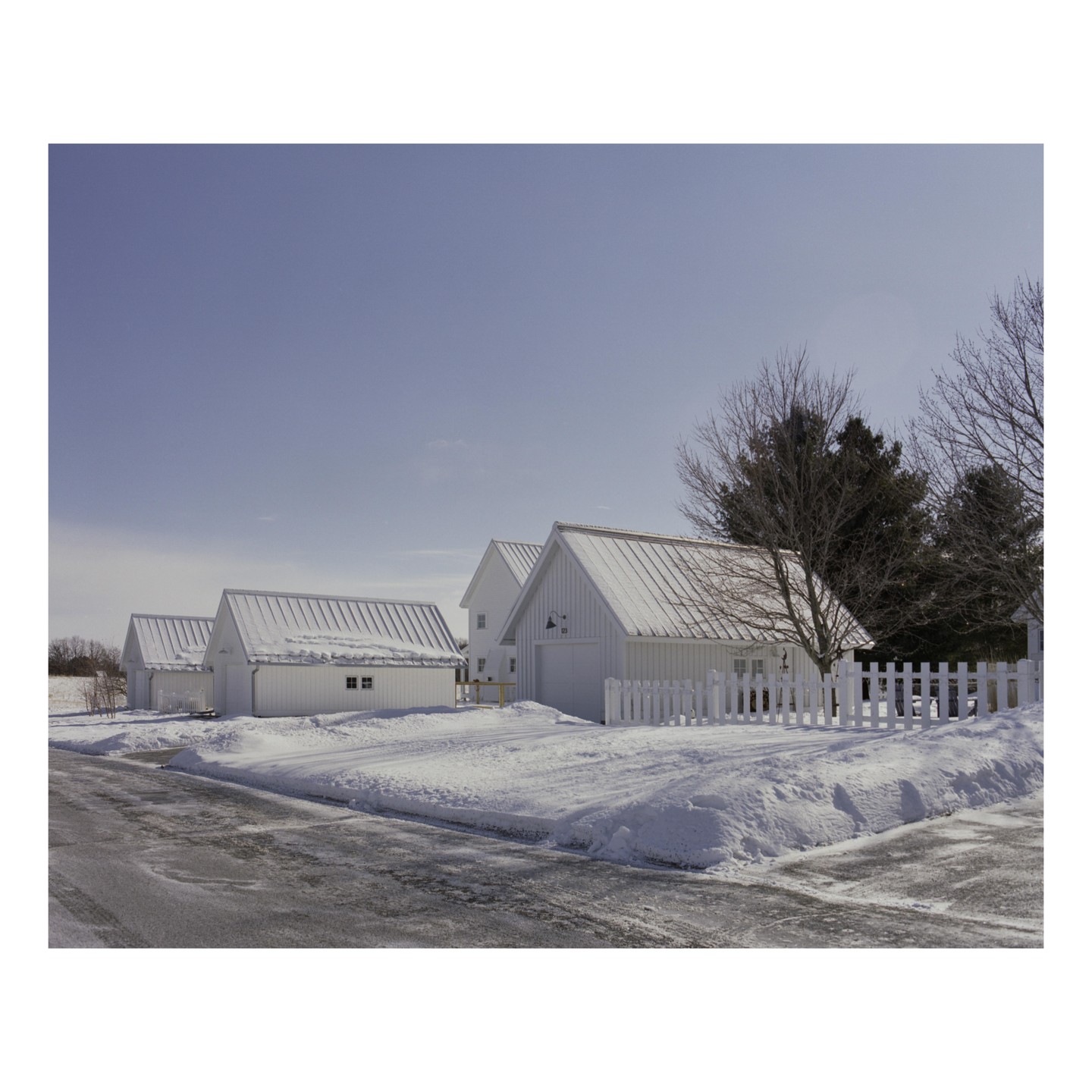 A row of white buildings with snow-covered roofs stands beside a road in a winter landscape.