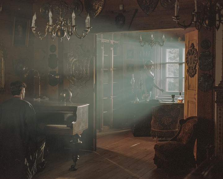 A man sits at a piano in a dimly lit room while a woman stands by a sunlit window in an adjacent room.