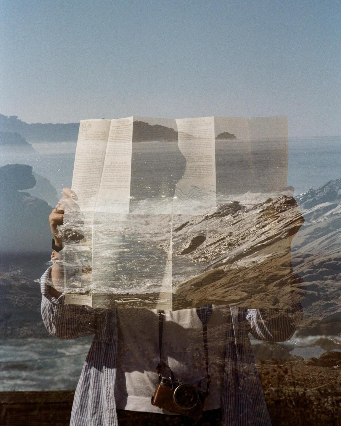 A person holds a map over a landscape with a body of water and mountains, creating a double exposure effect.