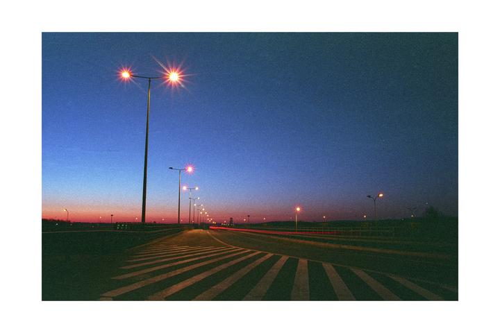 A long exposure captures light trails from vehicles on a road lined with streetlights at dusk.